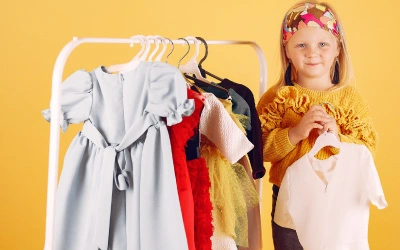  A kid is smiling, and a variety of kids clothes are hanging on a rack